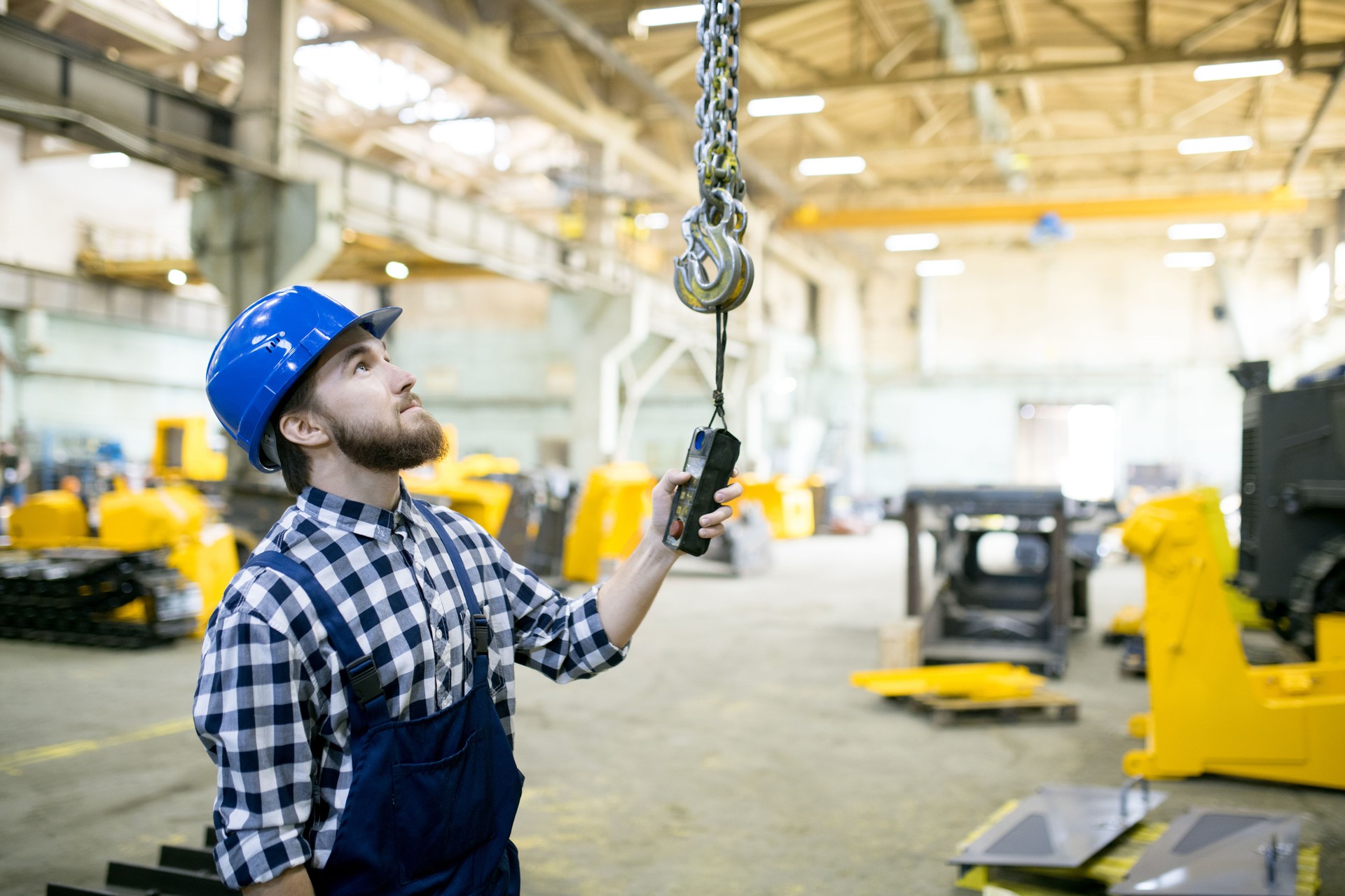 Skilled handsome young manual worker using overhead crane controller and pushing button while managing crane at industrial tractor plant shop